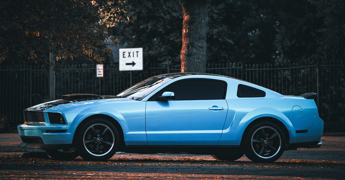 A classic blue Mustang coupe parked on a city street, highlighted by evening light and surrounded by trees.