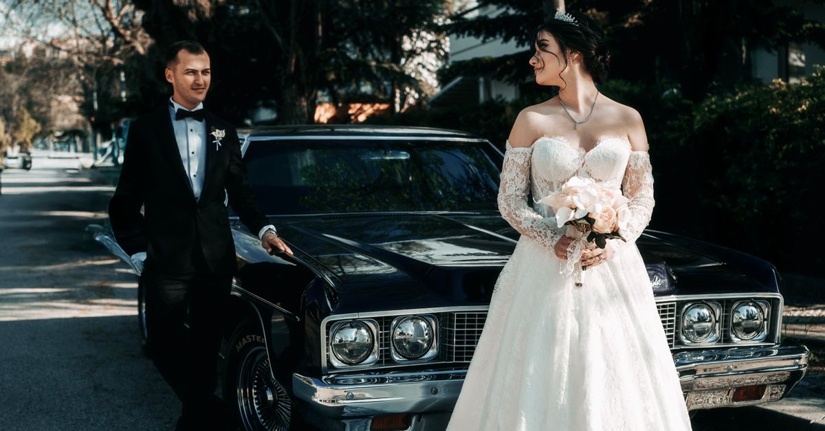 Bride and groom in wedding attire posing with vintage car on a sunny day.
