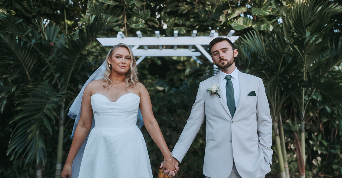 A bride and groom holding hands under a white arch in an outdoor wedding setting.
