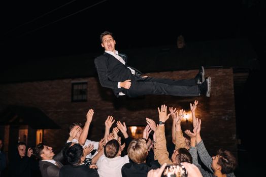 A lively wedding party scene with guests lifting a man in the air during the night celebration.