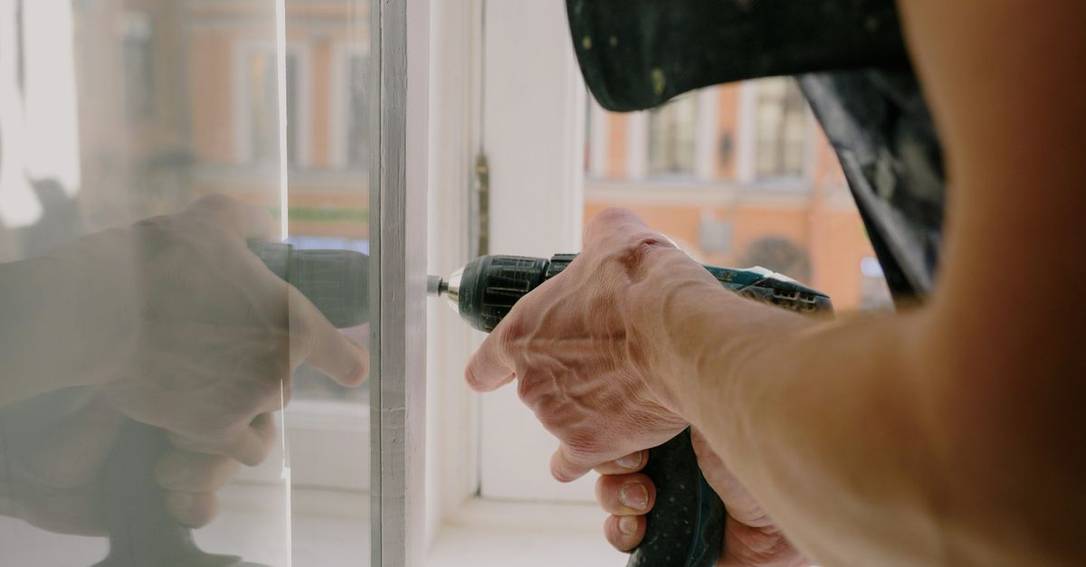 Unrecognizable professional male worker assembling white window with cordless screwdriver with drill near windowsill with reflection on glass during repair works in apartment