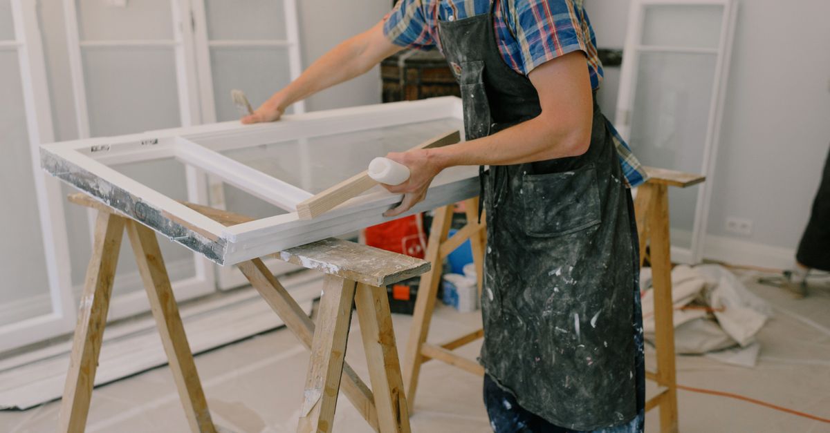 Unrecognizable male master wearing dirty apron standing near window with white frames on wooden workbench while working in professional studio